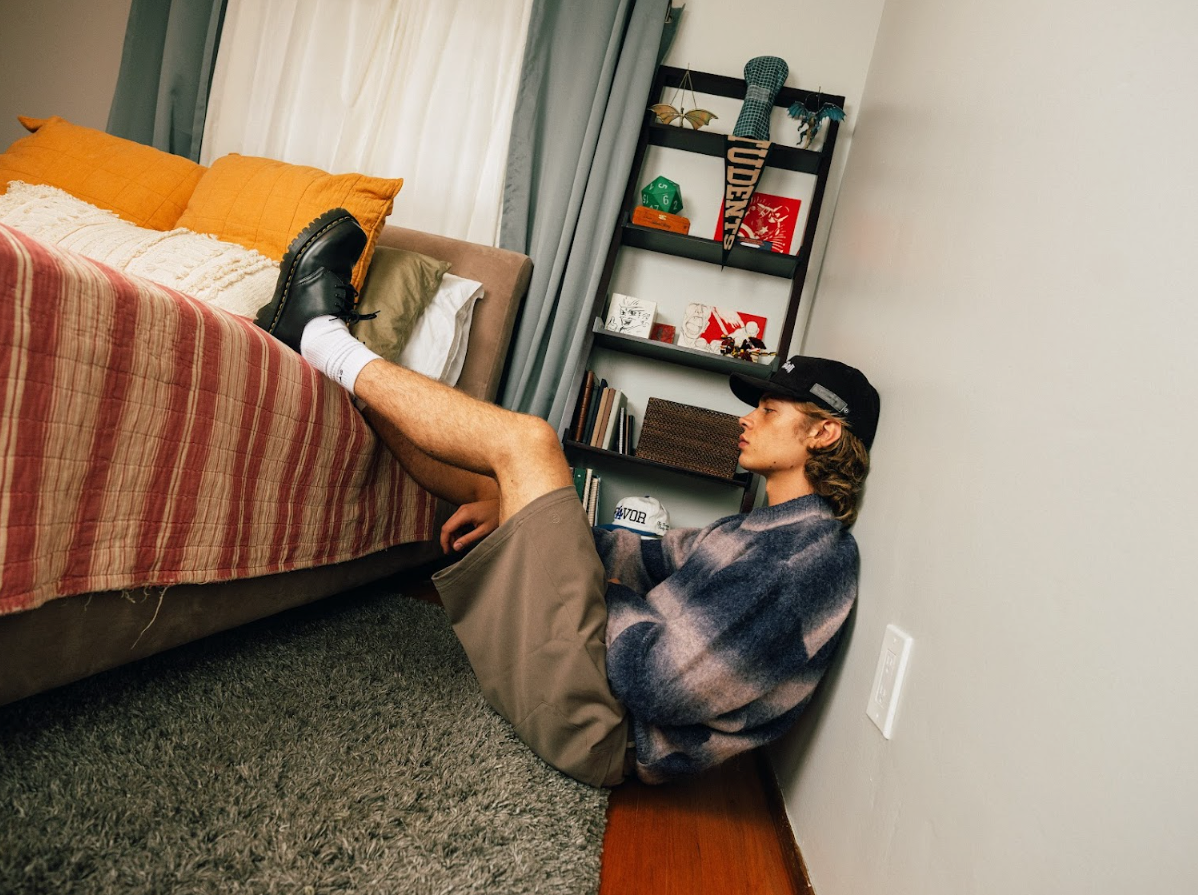 Person lying on the floor next to a bed in a room with a bookshelf.