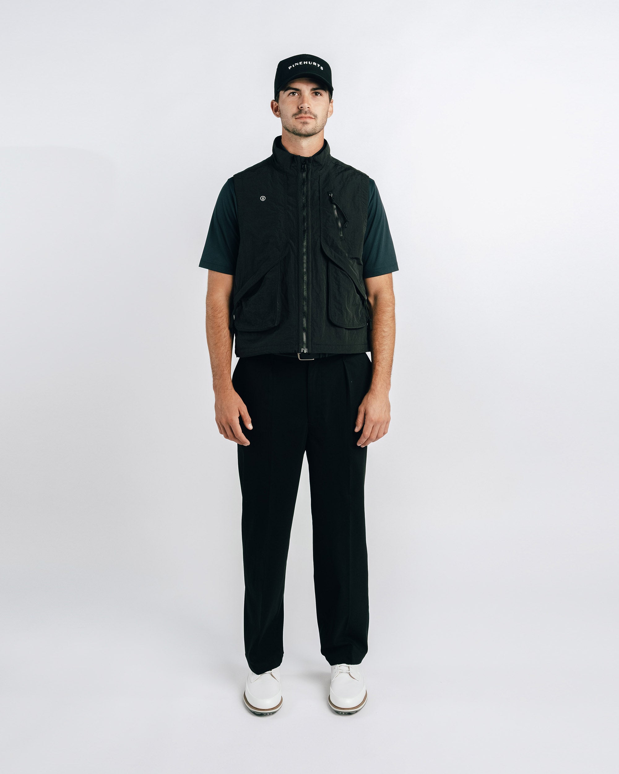 Man wearing a black vest over a dark shirt with black pants on a white background