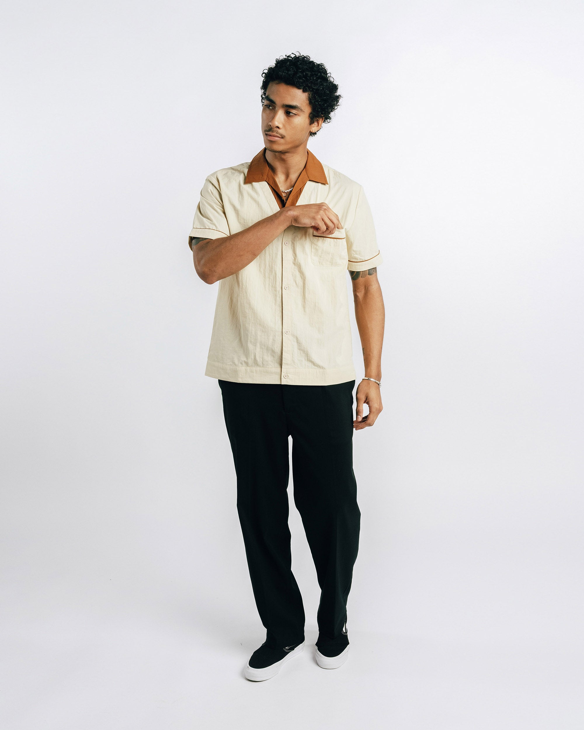 Man wearing a beige shirt with a brown collar and black pants on a white background