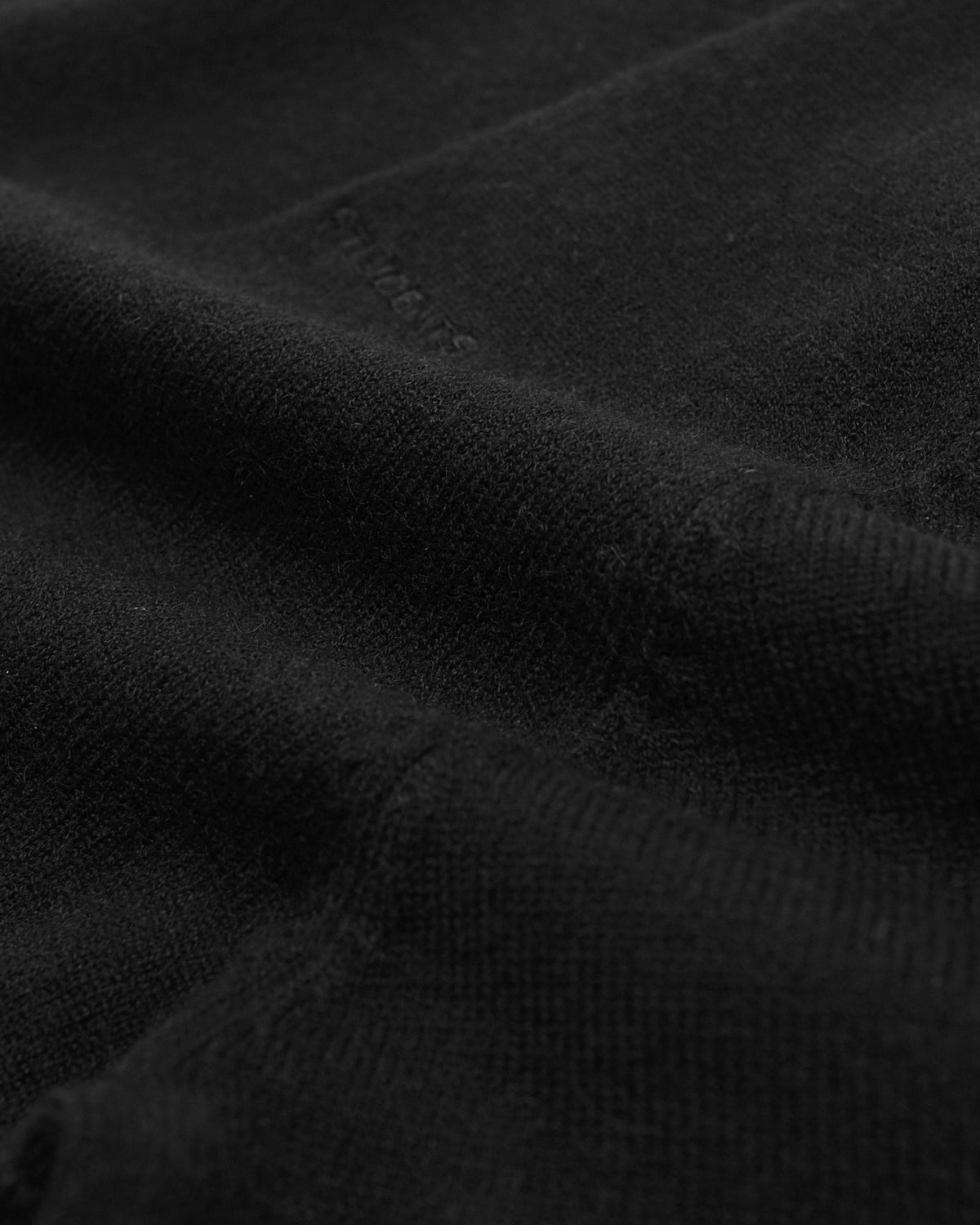 Close-up of a black knit fabric with a fine ribbed texture and a subtle tonal embroidered logo near the top.