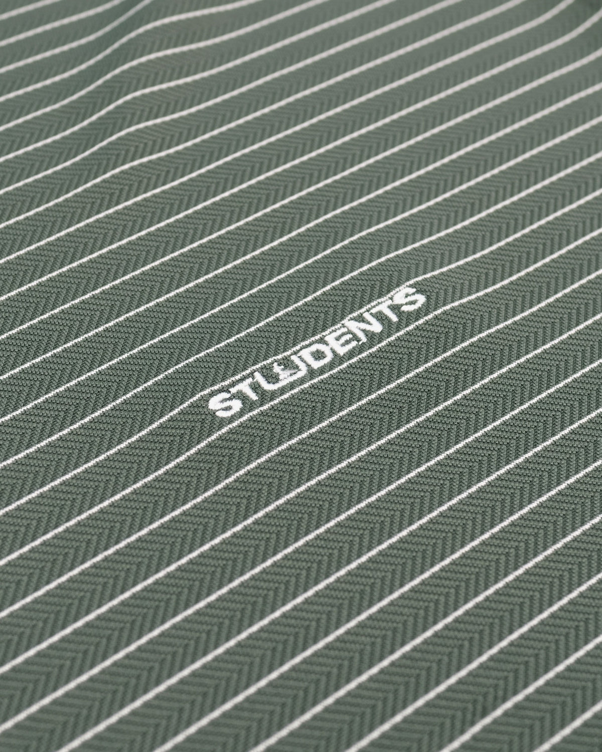 Close-up of a textured fabric with a dark green and gray herringbone pattern, featuring thin white diagonal stripes and the word 'STUDENTS' printed in white.
