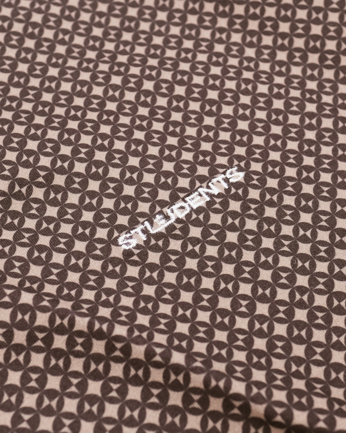 A close-up of a fabric surface with a repeating dark brown geometric pattern on a light beige background, featuring the word 'STUDENTS' embroidered in white near the center.