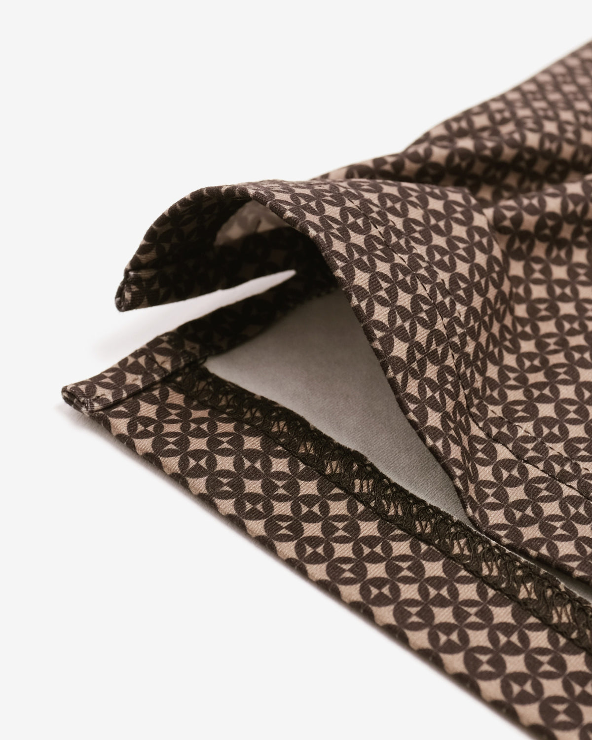 A close-up of a brown and beige patterned fabric, likely a tie or scarf, folded to show its woven geometric design and light gray interior lining, set against a plain white background.