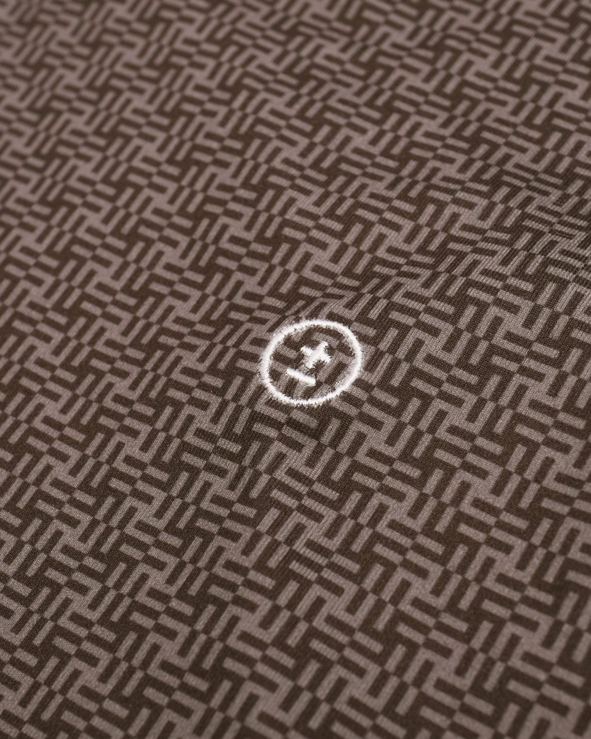 A close-up of a brown and beige geometric woven fabric pattern with a small white embroidered circular logo in the center.