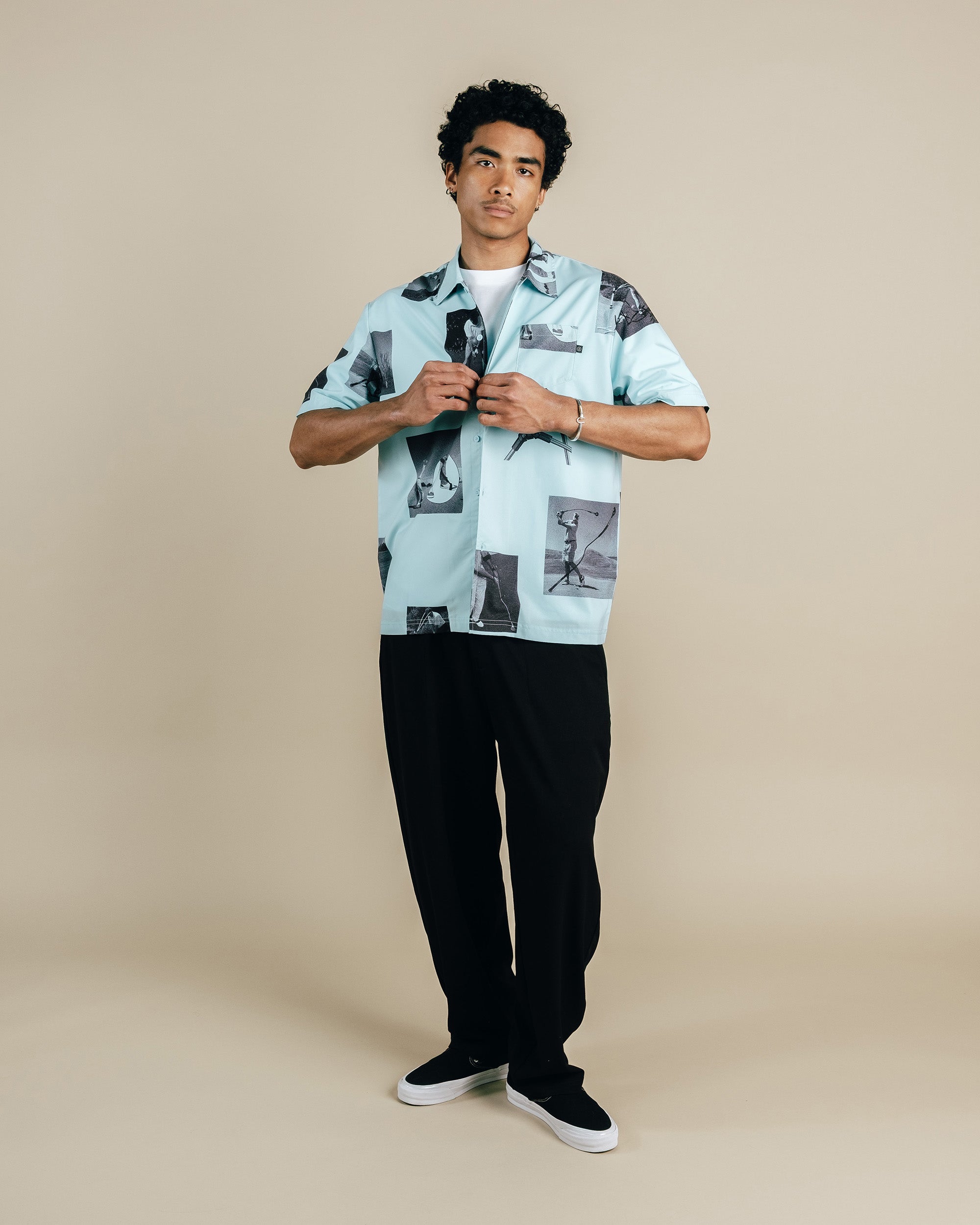 Man wearing a light blue patterned shirt and black pants on a beige background