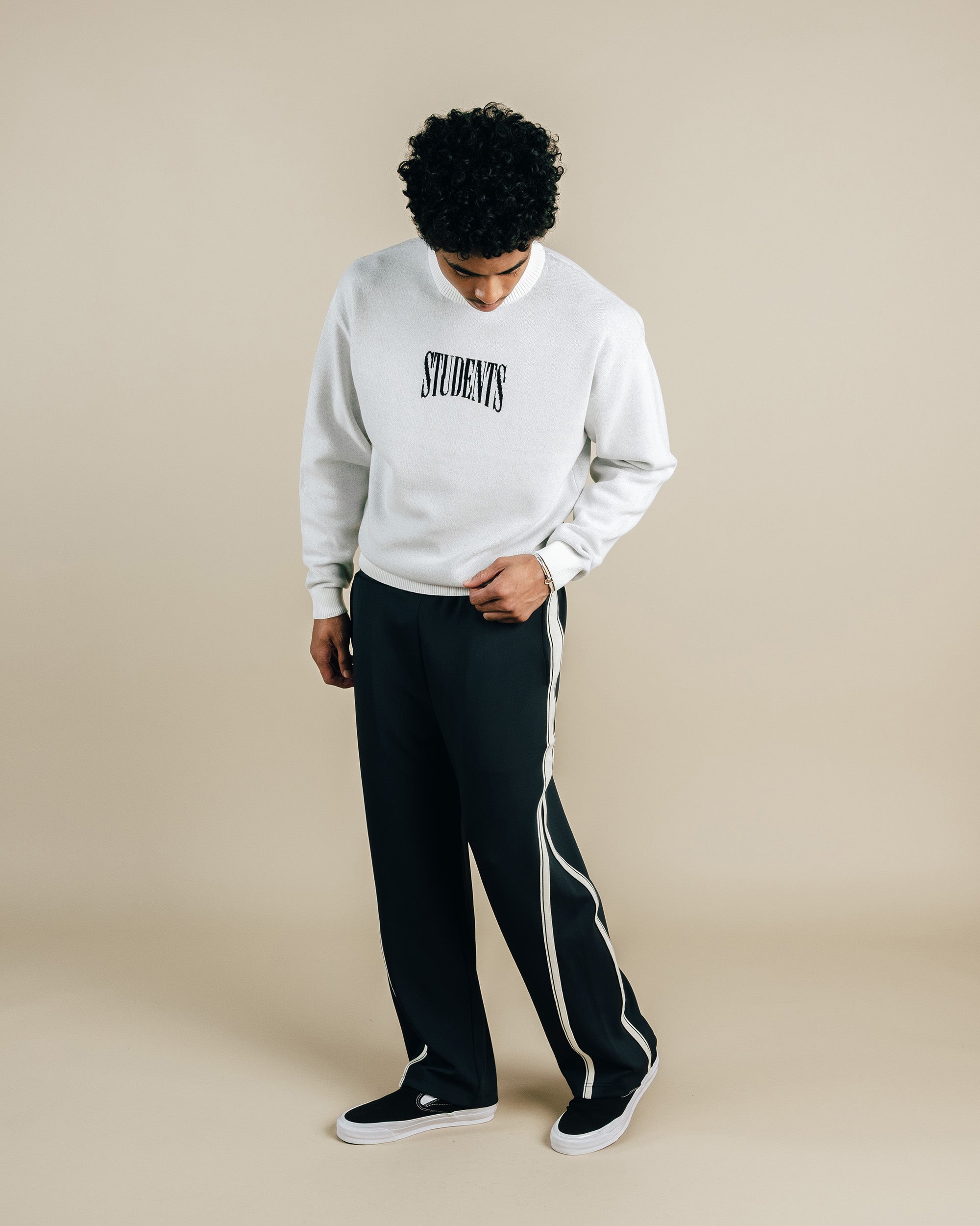 Person wearing a white sweatshirt with 'STUDENTS' text and black pants with white stripes on a beige background