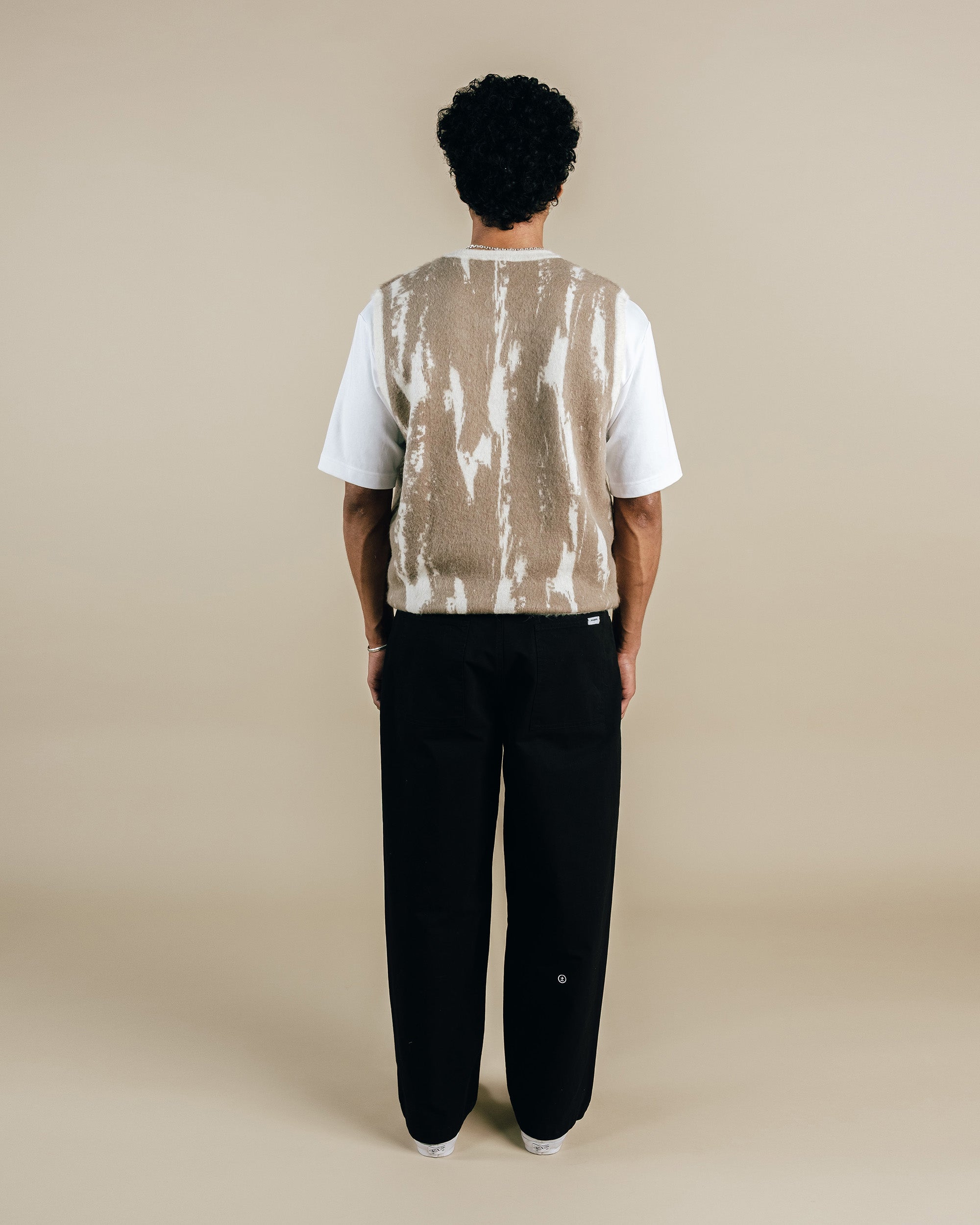Person wearing a white shirt, beige vest, and black pants on a beige background