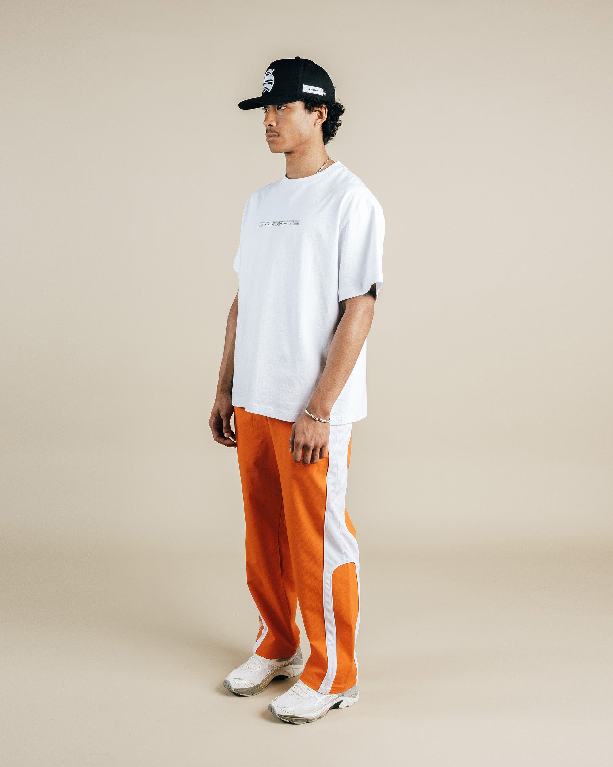 Person wearing a white t-shirt, orange pants, and a black cap on a beige background