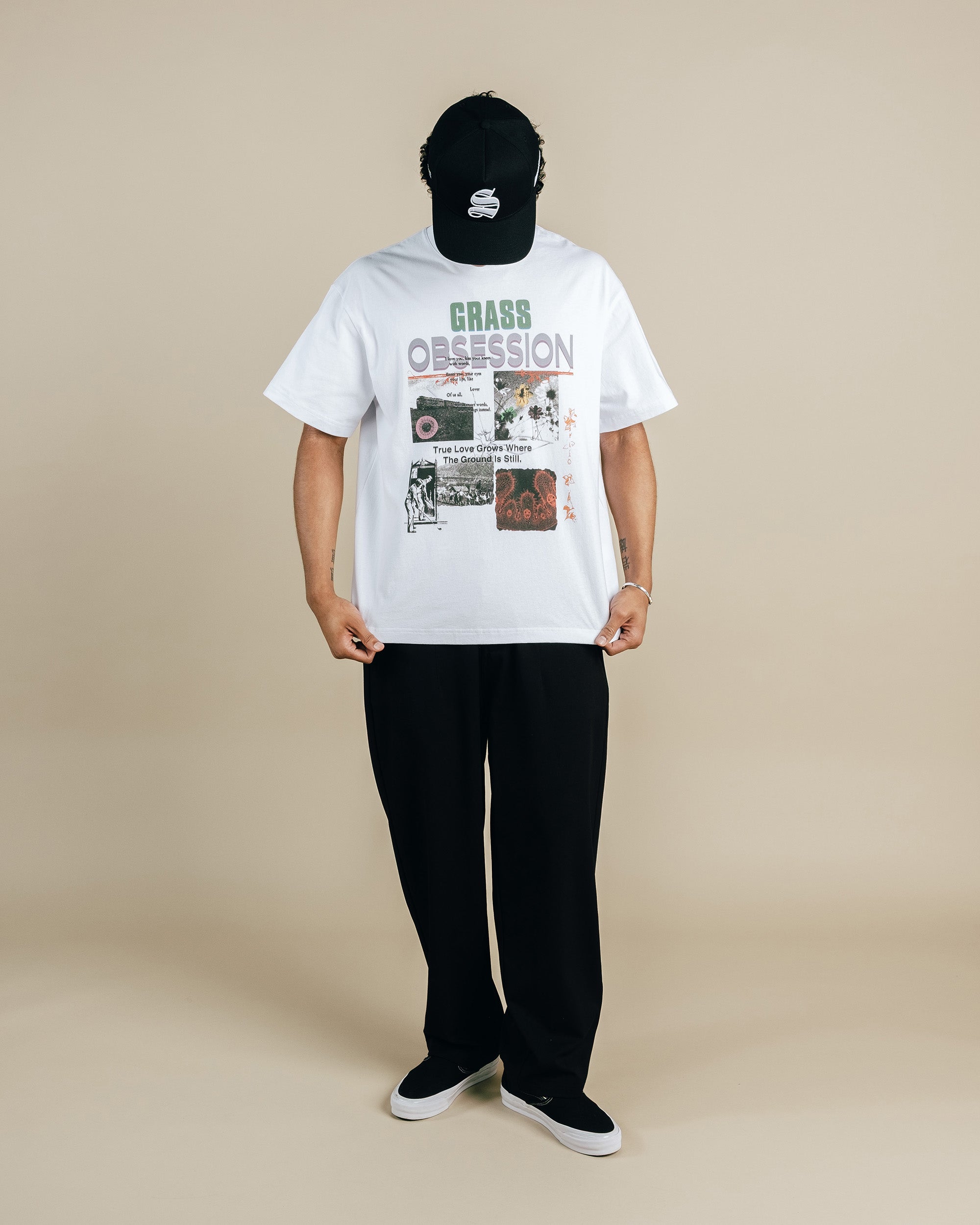 Person wearing a white t-shirt with graphic design and text, black pants, and a black cap on a beige background