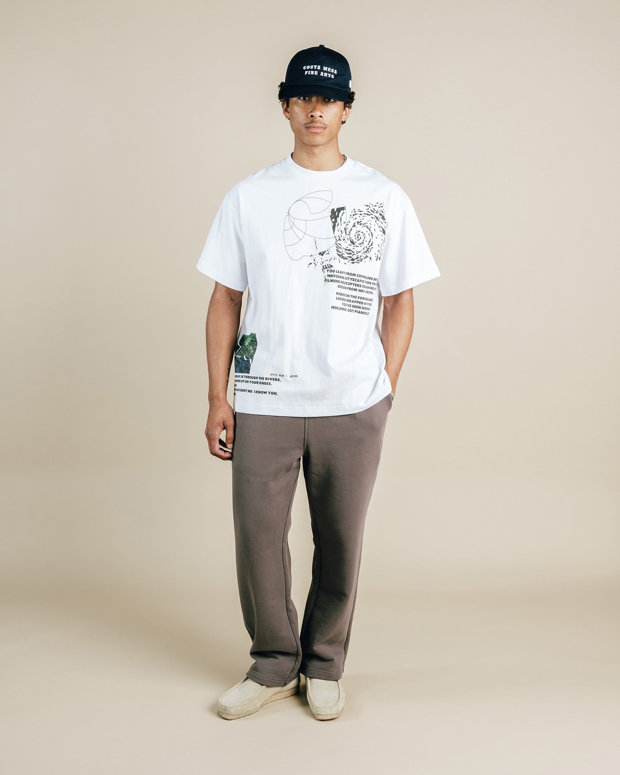 Person wearing a white t-shirt with graphics and text, gray pants, and a black cap on a beige background