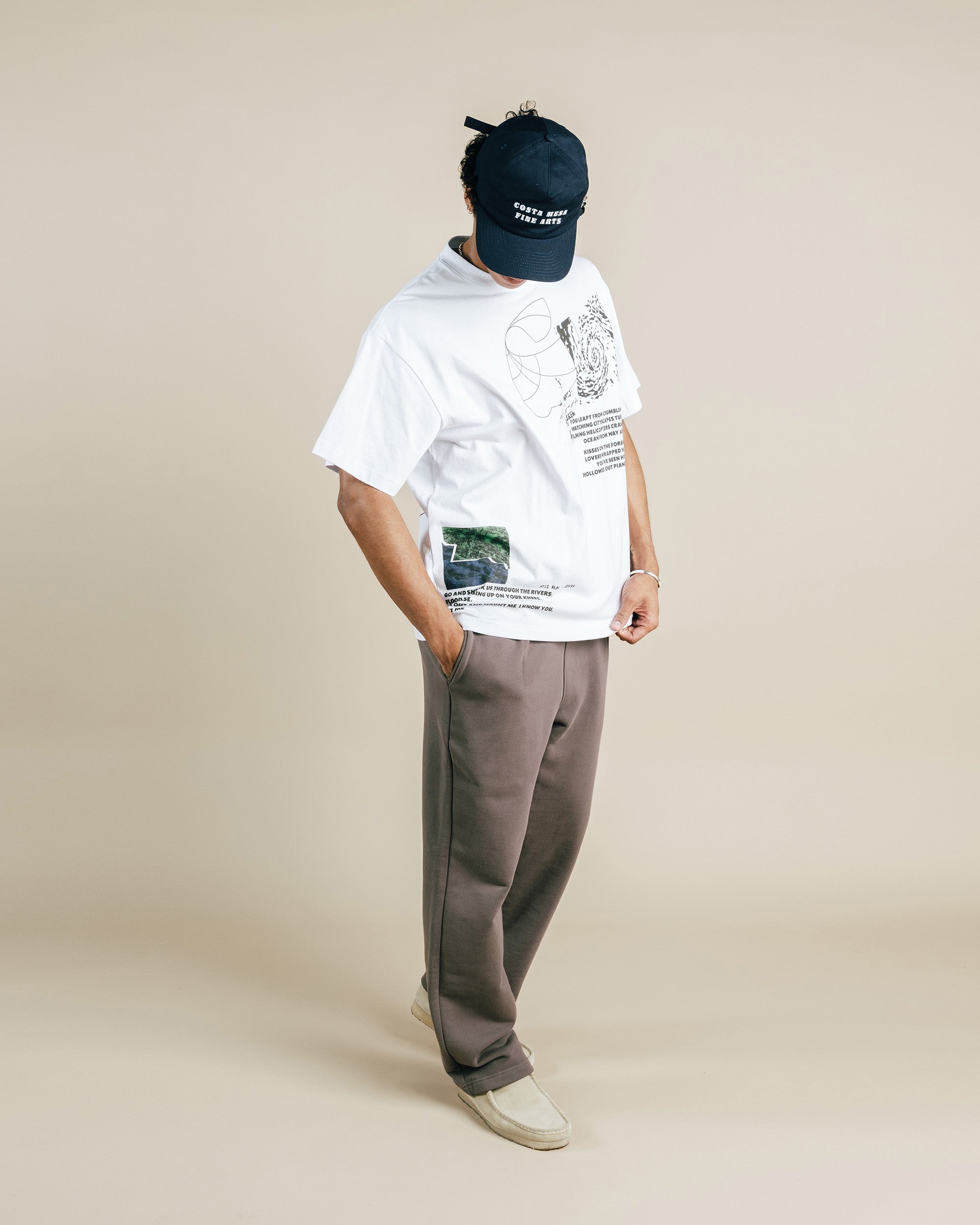 Person wearing a white t-shirt with graphics and text, gray pants, and a dark cap on a beige background