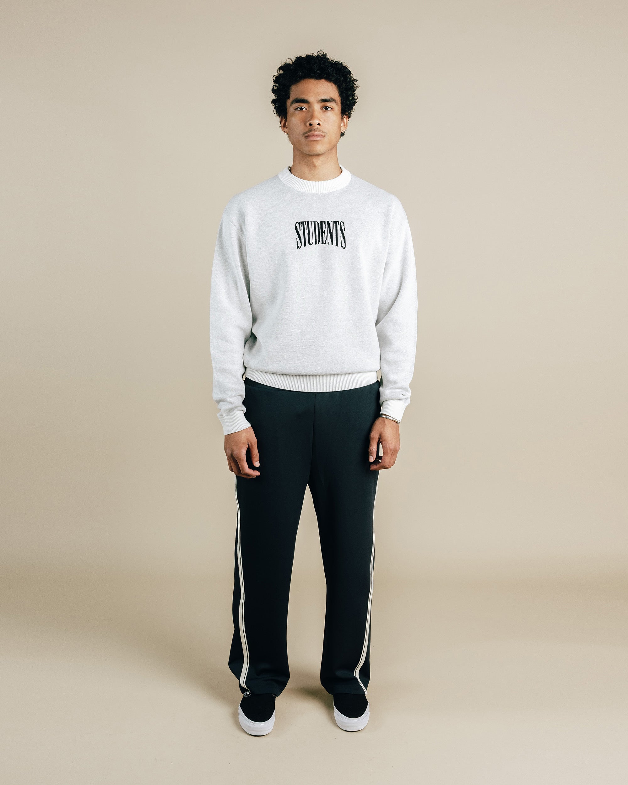 Person wearing a white sweatshirt with 'STUDIORS' text and black pants with white stripes on a beige background