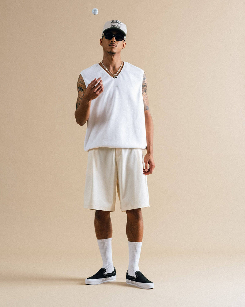 Person wearing a white sleeveless shirt, beige shorts, black sneakers, and a cap on a beige background