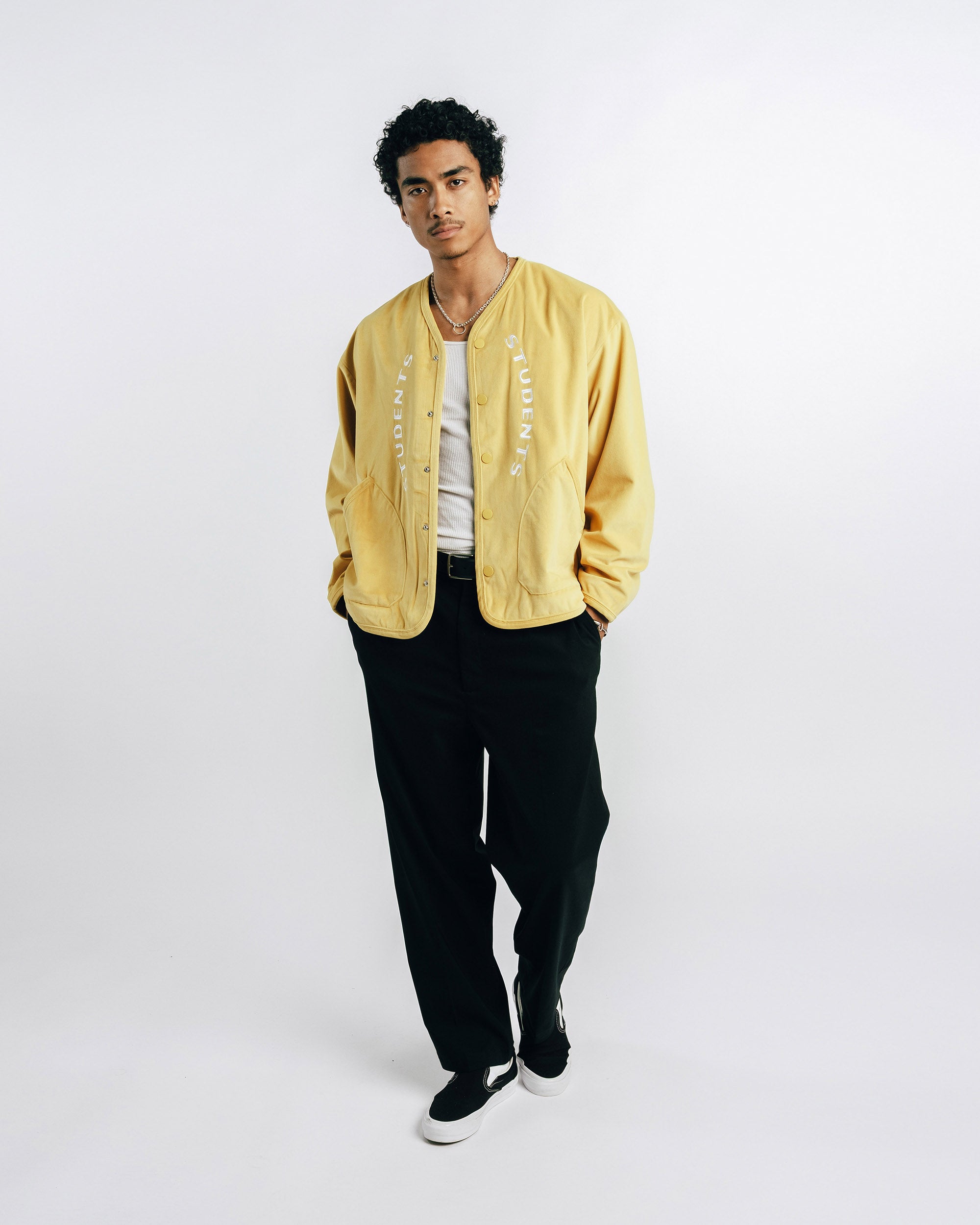 Person wearing a mustard yellow jacket, white shirt, black pants, and black shoes on a white background