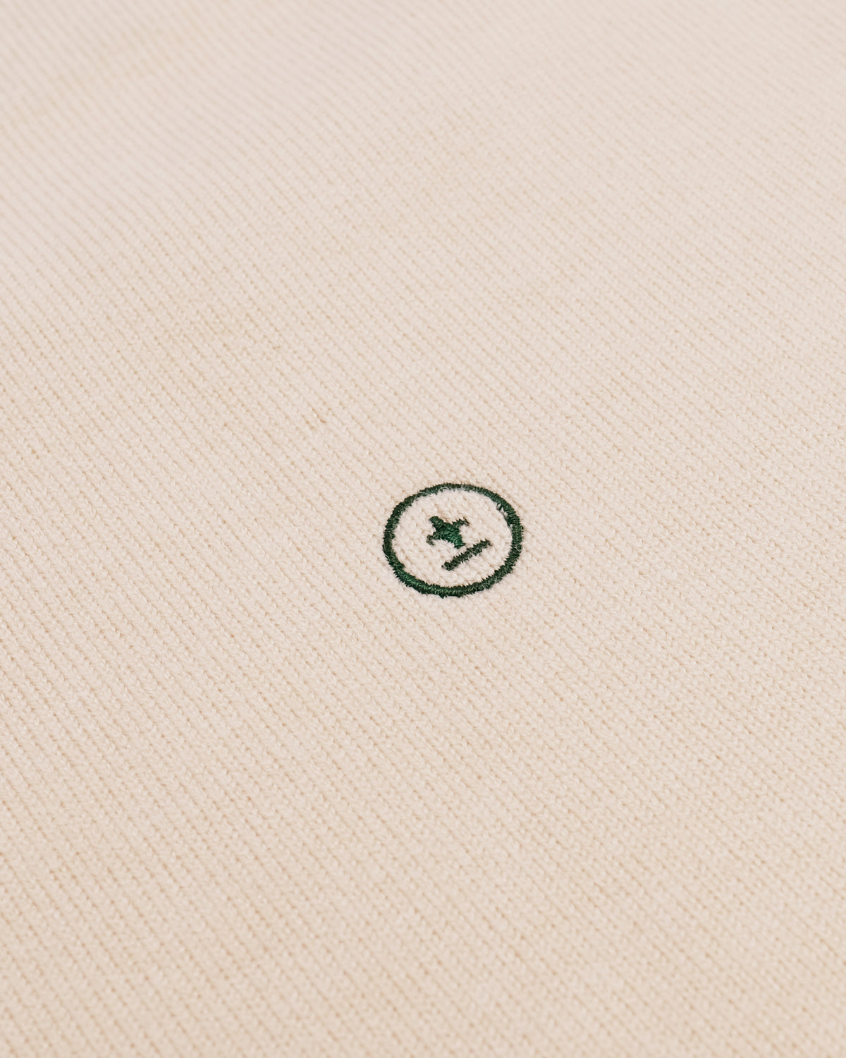 Close-up of a cream-colored textured fabric with a small dark green embroidered circular logo featuring a star and diagonal line.