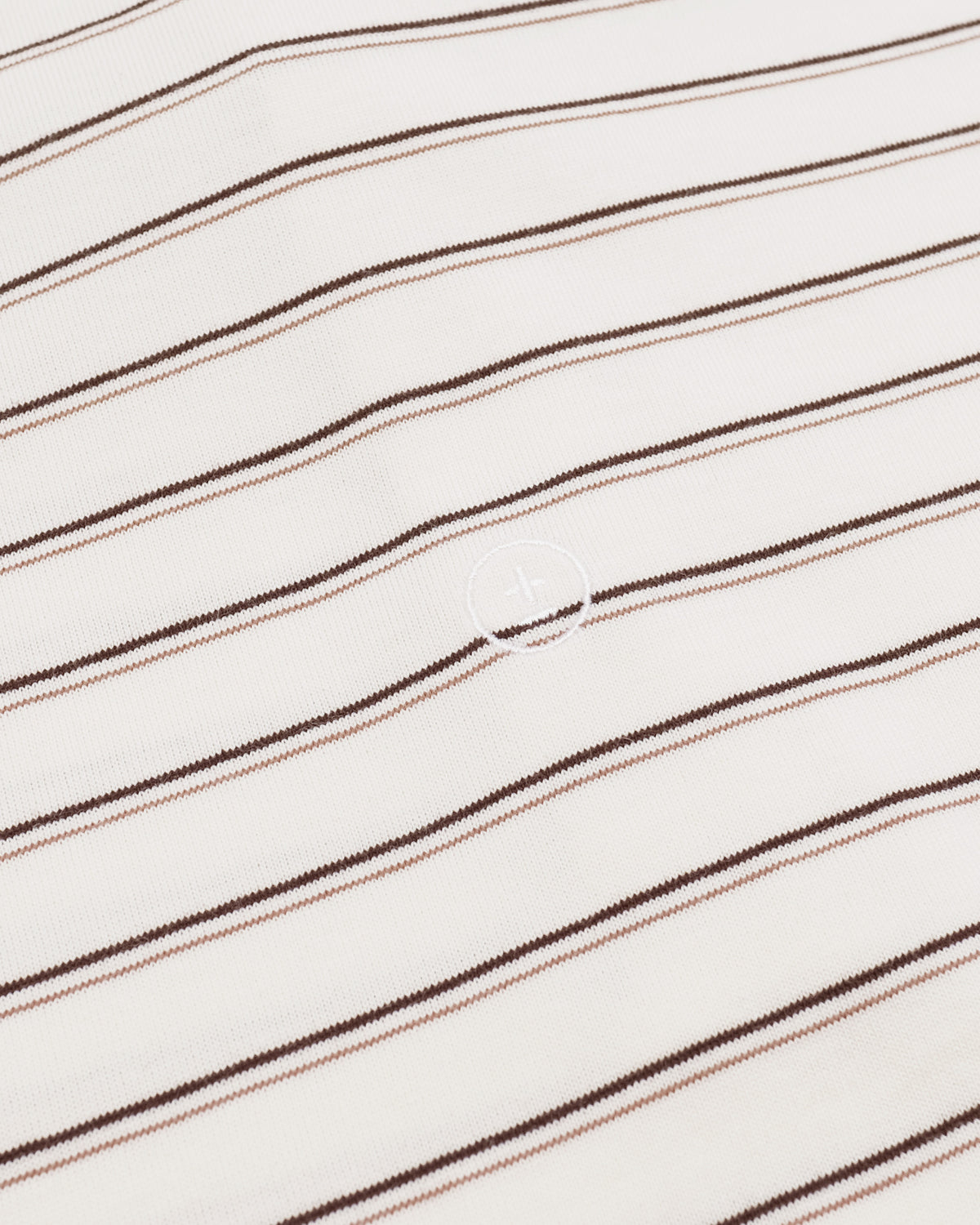 A close-up of a white or off-white knit fabric with thin diagonal stripes in dark brown and a muted dusty pink, featuring a small circular logo in the center.
