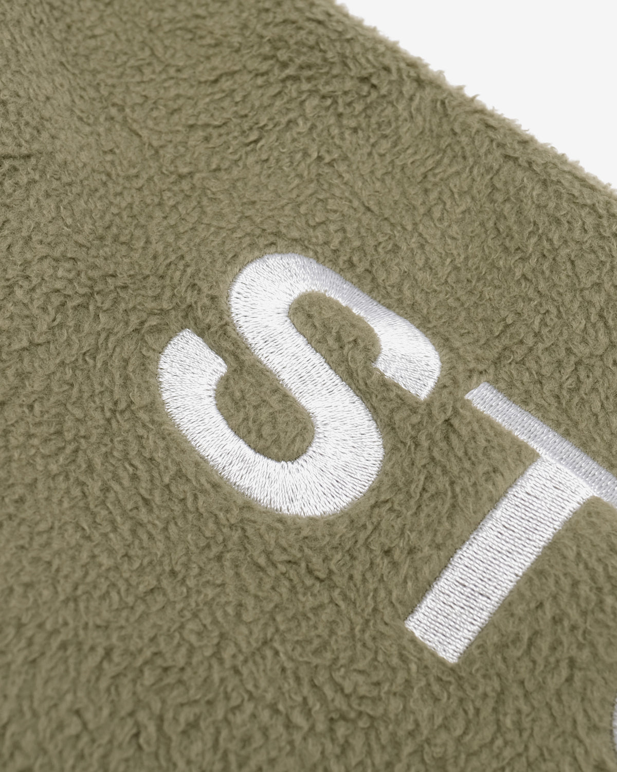 Close-up of a textured olive green fleece or sherpa fabric with a large white embroidered letter 'S' and part of another white letter visible.
