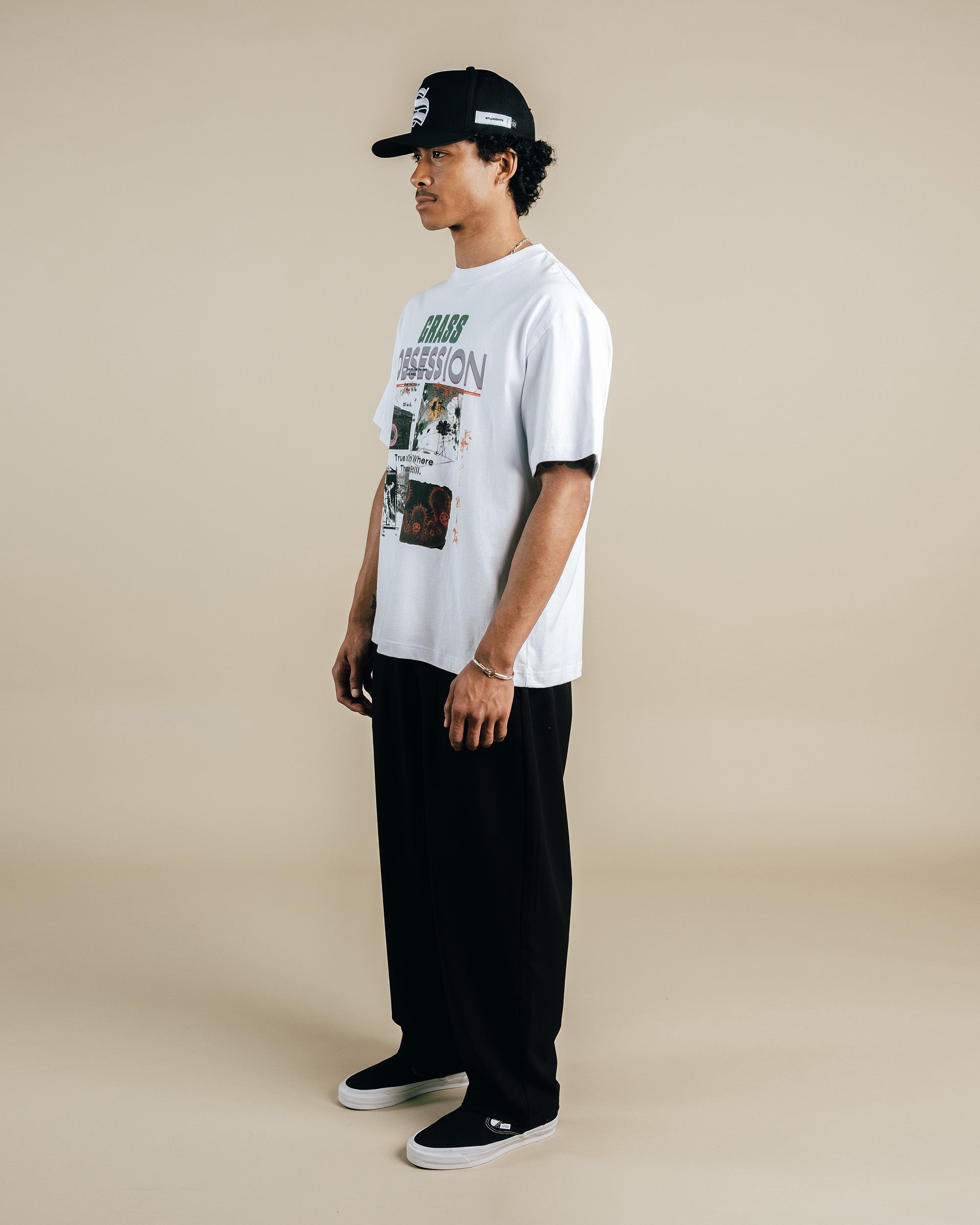 Person wearing a white graphic t-shirt, black pants, and a black cap on a beige background