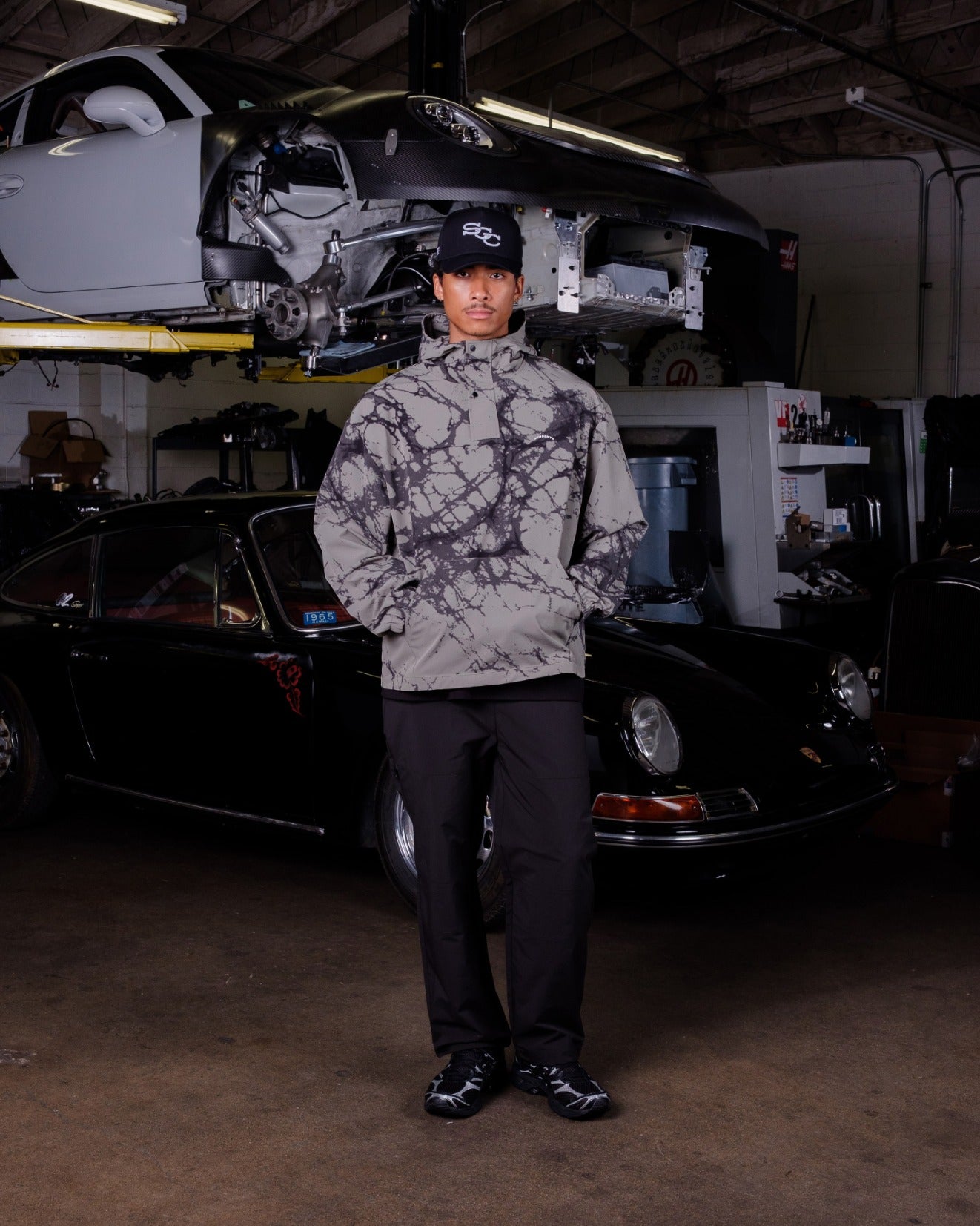 Person wearing a camouflage jacket and cap in a garage with cars and equipment.
