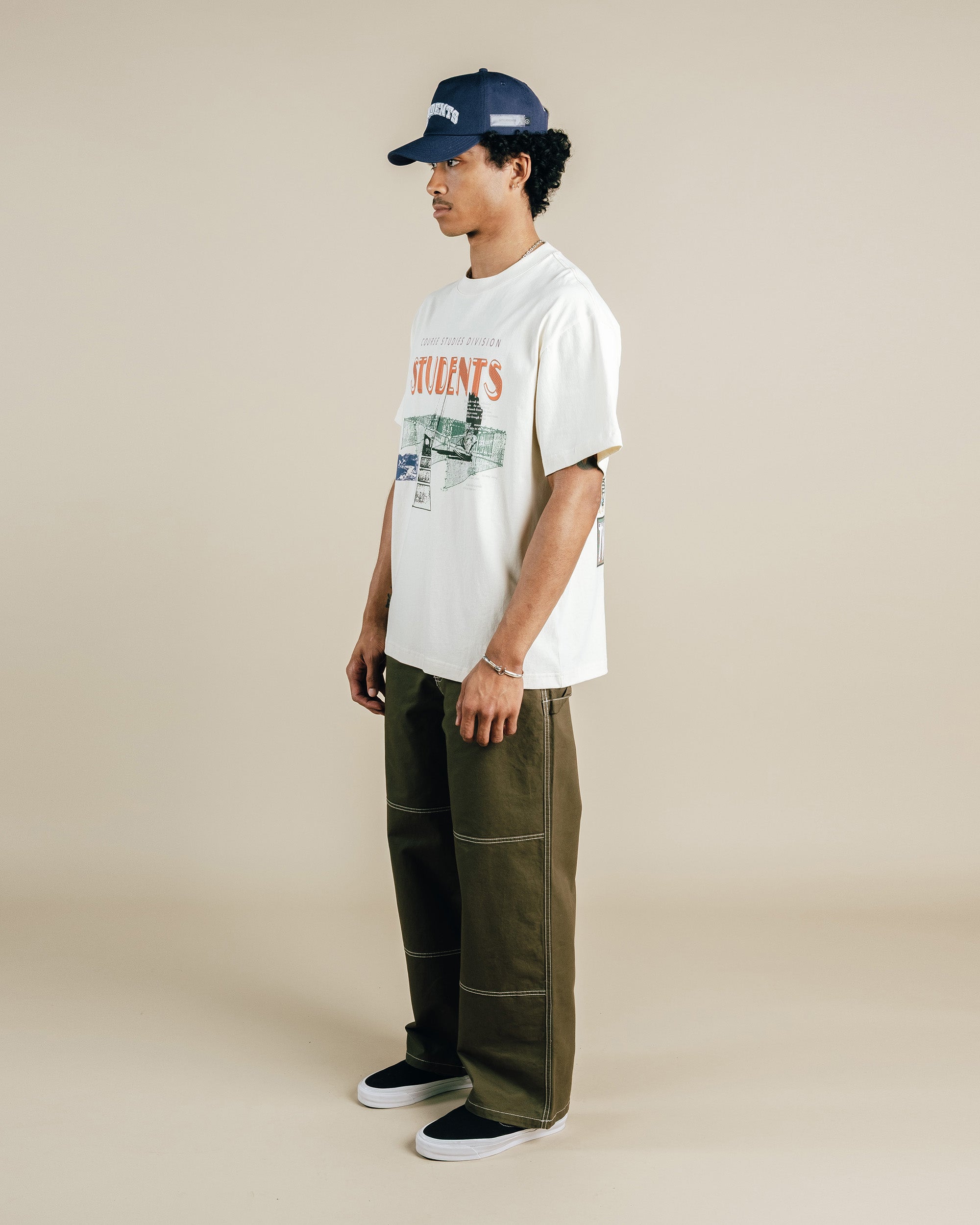 Person wearing a nude t-shirt with graphics, green pants, and a navy cap on a beige background