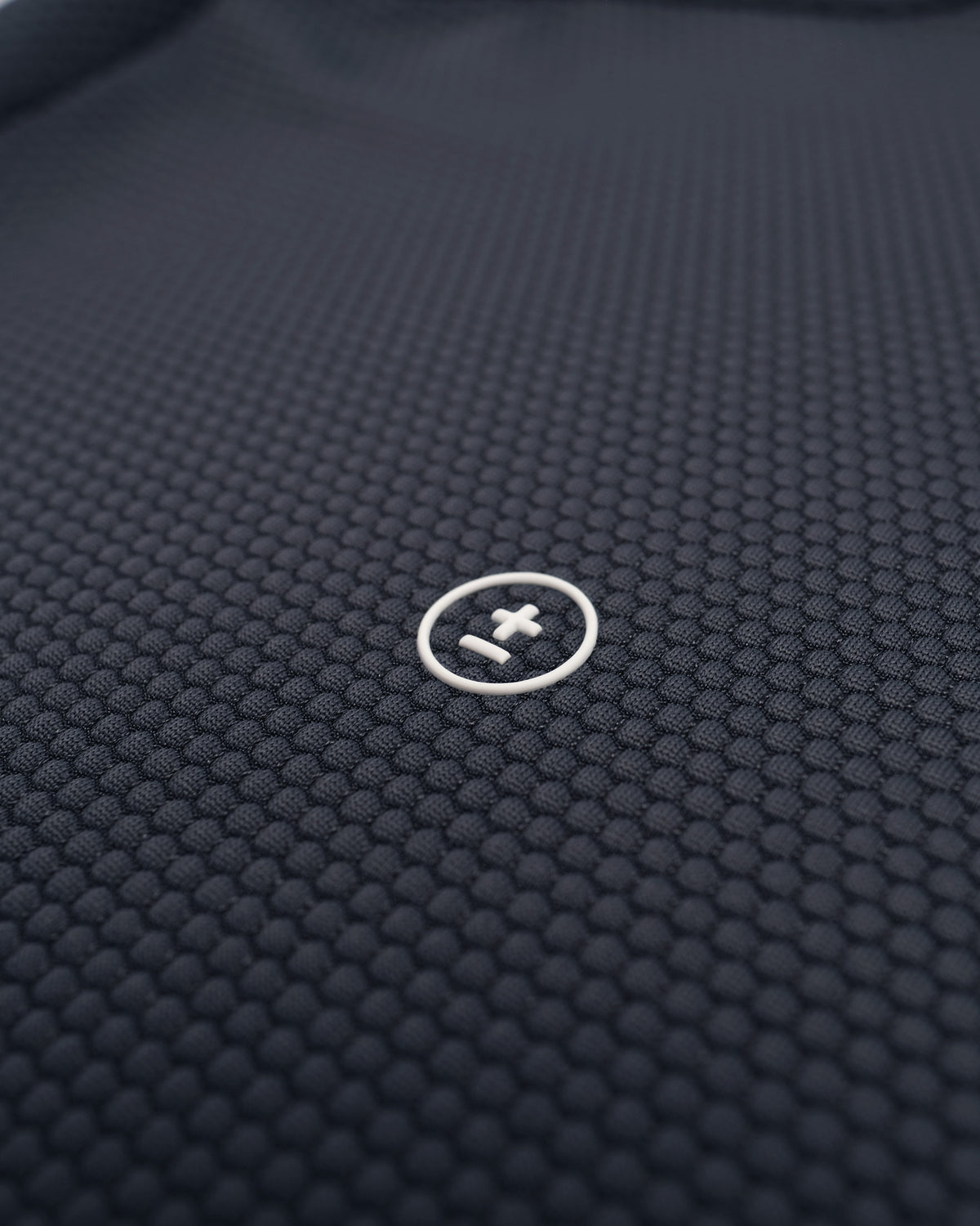 Close-up of a dark navy textured athletic fabric with a small circular white logo featuring a plus-minus symbol.